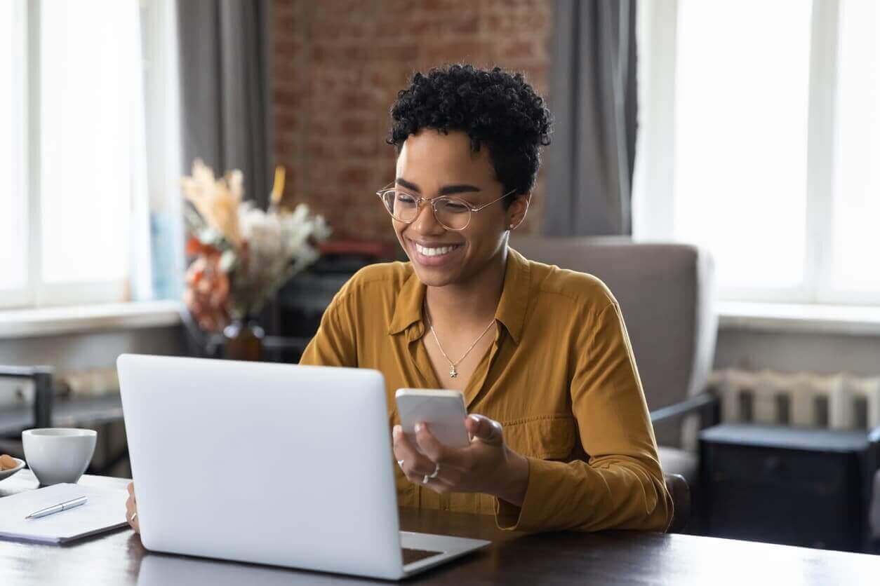 African woman sit at desk holds cellphone staring at laptop